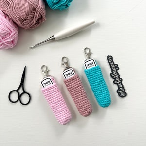 May include: Three crocheted lip balm holders in pink, light brown, and turquoise. Each holder has a white label that reads "Lip Balm Holder". The holders are displayed on a white surface with yarn balls and a crochet hook.