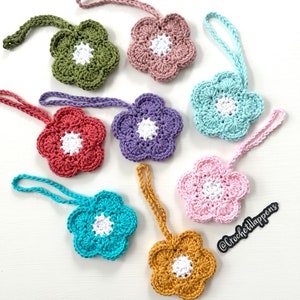 May include: Seven crocheted flower ornaments in various colors, including green, pink, blue, purple, red, and yellow. Each flower has a white center and a loop for hanging. The flowers are arranged on a white surface. The text "@crochet.happens" is written in black on a white background in the lower right corner of the image.