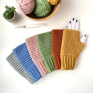 May include: A set of six fingerless gloves in gray, blue, pink, green, rust, and mustard yellow, each featuring a ribbed cuff and a textured crochet stitch. A wooden bowl filled with matching yarn balls and a crochet hook are positioned nearby.