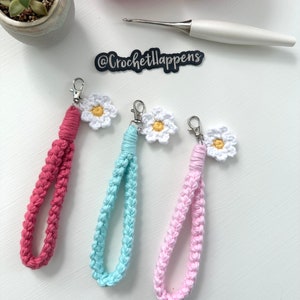 May include: Three crocheted keychains with daisy charms. The keychains are pink, blue, and red. The charms are white with yellow centers. The image also includes a crochet hook and balls of yarn.