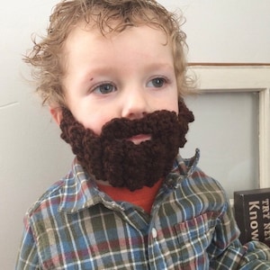 May include: A young child wearing a brown crocheted beard and a plaid shirt.