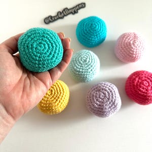 May include: A hand holding a teal crocheted ball, surrounded by other colorful crocheted balls in shades of blue, pink, yellow, and purple. The balls are arranged on a white surface. The text "@crochetlhappens" is visible.