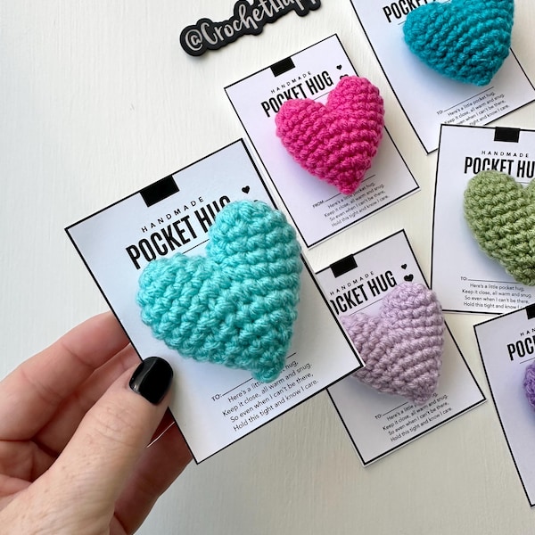 Peut inclure: Des câlins de poche en forme de cœur en crochet faits à la main dans différentes couleurs, notamment turquoise, rose, lavande, vert et violet. Chaque cœur est accompagné d'une étiquette qui indique "Câlin de poche fait main" et comprend un message : "Voici un petit câlin. Garde-le près de toi, bien au chaud et bien serré. Même quand je ne suis pas là, serre-le fort et sache que je pense à toi."