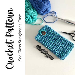 May include: A handmade crochet sunglasses case in shades of blue and teal, secured with a wooden button. The text "Crochet Pattern Sea Glass Sunglasses Case" is visible. Yarn, a crochet hook, and scissors are also present.