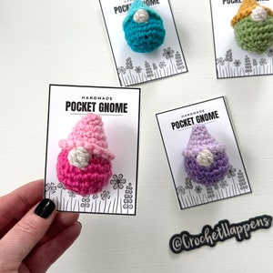 May include: Four handmade crocheted gnome ornaments on white cards with black text that reads "Handmade Pocket Gnome". The gnomes are pink, purple, teal, and green. Each gnome has a white face and a white hat.