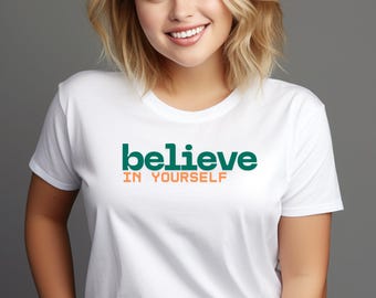 May include: White t-shirt with the words "believe IN YOURSELF" printed in green and orange. The shirt has a crew neck and short sleeves. The text is centered on the chest.