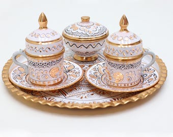 May include: A decorative coffee set featuring two cups with lids, saucers, and a sugar bowl, all resting on a gold-rimmed tray. The set is white with gold and black floral patterns. The cups have small handles and gold accents.
