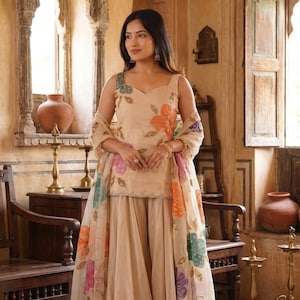May include: A beige sleeveless top and wide-leg pants set featuring floral embroidery in orange, pink, green, and purple. A matching sheer dupatta with similar floral embroidery is draped over the shoulders. The text Krishna Saree Shop is visible.