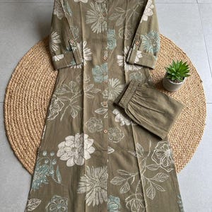 Puede incluir: Conjunto de túnica y pantalón con estampado floral verde oliva. La túnica presenta una botonadura frontal, mangas tres cuartos y cuello mandarín. El diseño floral incluye detalles en blanco y turquesa. El pantalón tiene cintura elástica.
