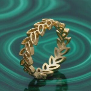 May include: A gold ring with a repeating leaf pattern. The ring's leaves are finely detailed, giving it a delicate, nature-inspired look. The ring is set against a green background.