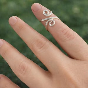 May include: A silver-colored ring with a spiral design. The ring wraps around the finger with two swirling accents. The ring is worn on a finger against a blurred green background.