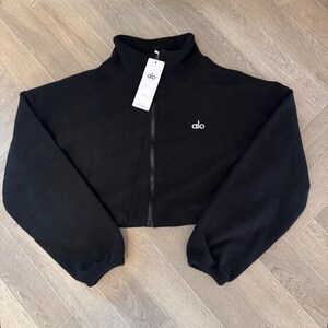 May include: A black cropped zip-up jacket with a high collar and long sleeves featuring elastic cuffs. The garment has a central black zipper, the white text alo printed on the left chest, and a white rectangular brand tag attached to the collar.