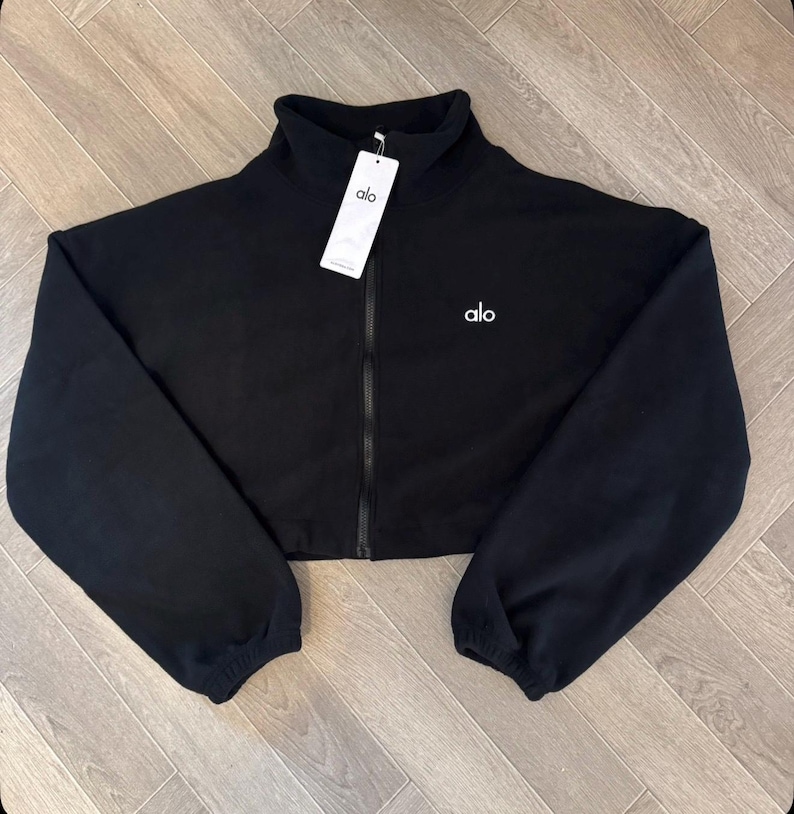 May include: A black cropped zip-up jacket with a high collar and long sleeves featuring elastic cuffs. The garment has a central black zipper, the white text alo printed on the left chest, and a white rectangular brand tag attached to the collar.