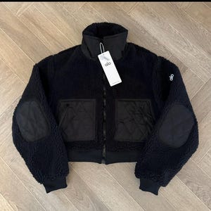 May include: A black Alo brand jacket made of textured fleece material. The jacket features a high collar, long sleeves with quilted elbow patches, and two front pockets with quilted detailing. A white brand tag is attached to the collar.