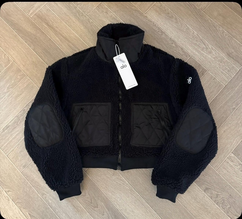 May include: A black Alo brand jacket made of textured fleece material. The jacket features a high collar, long sleeves with quilted elbow patches, and two front pockets with quilted detailing. A white brand tag is attached to the collar.