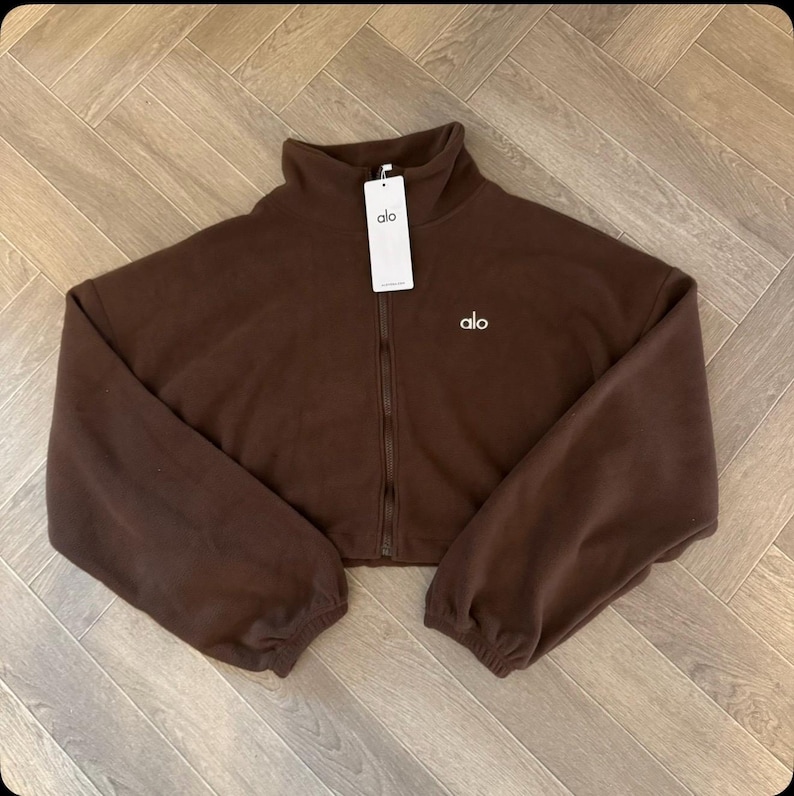 May include: A dark brown cropped fleece jacket with a high collar and a full-length front zipper. The jacket features long sleeves with elastic cuffs and the white text 'alo' embroidered on the left chest, with a matching white brand tag attached.