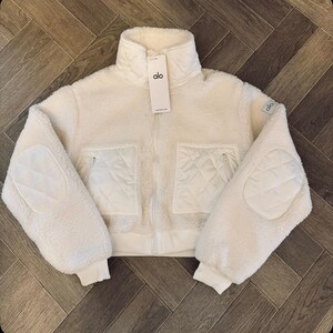 May include: A cream-colored, zip-up sherpa jacket featuring a high collar, long sleeves with quilted elbow patches, and two front pockets with quilted detailing. The jacket includes a white brand tag labeled alo hanging from the zipper pull.
