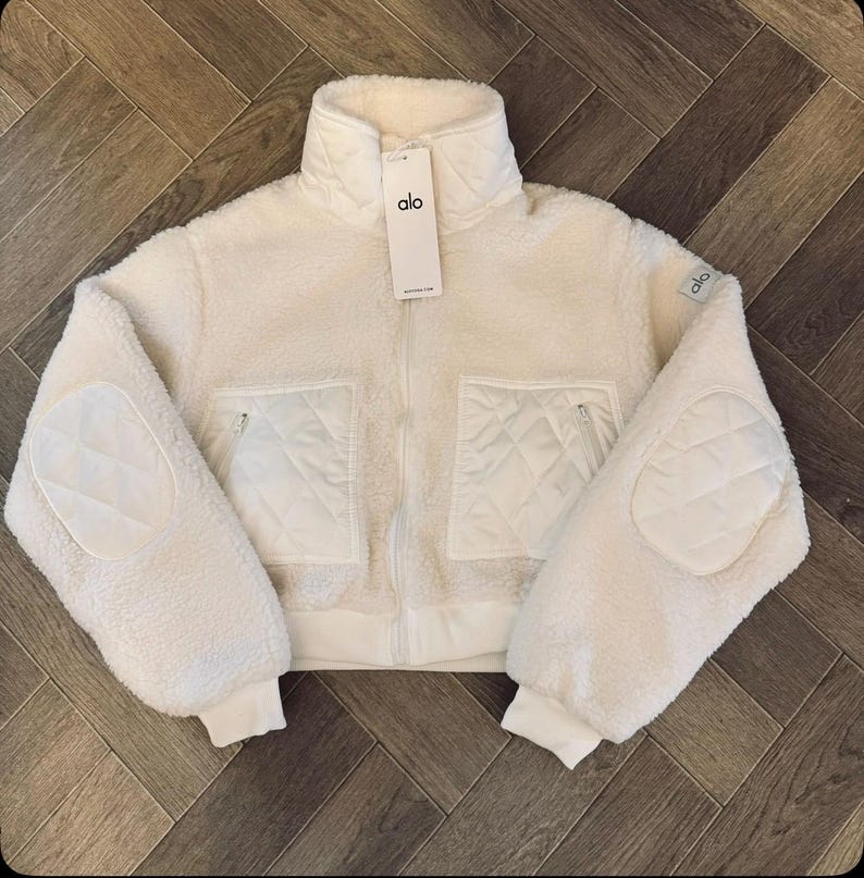 May include: A cream-colored, zip-up sherpa jacket featuring a high collar, long sleeves with quilted elbow patches, and two front pockets with quilted detailing. The jacket includes a white brand tag labeled alo hanging from the zipper pull.