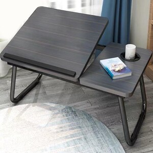May include: A dark grey wood-grain laptop desk with a foldable metal frame. The desk features an adjustable tilting surface with a black ledge, a stationary side section with a built-in circular cup holder, a white mug, and a small book.
