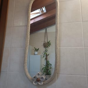 Peut inclure: Miroir ovale avec un cadre enroulé de corde. Le miroir est suspendu par une corde et comporte une fleur décorative en bas. Le miroir est accroché à un mur carrelé.