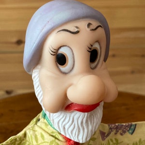 Puede incluir: Un títere vintage con un sombrero morado, una barba blanca y una nariz grande y redonda. El títere tiene una cara pintada con ojos grandes y expresivos.
