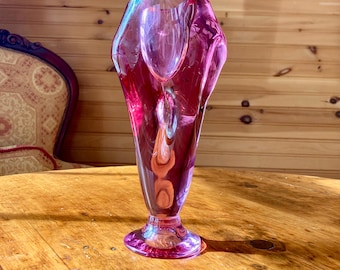 Può includere: Un vaso di vetro rosa con un design unico e astratto. Il vaso ha un'apertura larga e svasata e una base stretta.