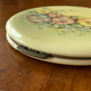Op de afbeelding: Een vintage compact spiegel met een bloemenmotief. De spiegel is ovaalvormig en heeft een witte achtergrond met een bloemenpatroon in roze, gele en blauwe tinten. De spiegel is gesloten en het scharnier is zichtbaar.