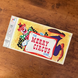 Puede incluir: Una caja de juego de mesa vintage con un payaso de dibujos animados sosteniendo un cartel que dice "The Merry Circus Game". La caja es amarilla con detalles rojos, azules y verdes.