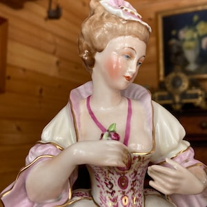 Puede incluir: Una figurita de porcelana de una mujer vestida con un vestido rosa y blanco con ribetes dorados. Tiene una flor rosa en la mano y lleva un sombrero rosa y dorado.