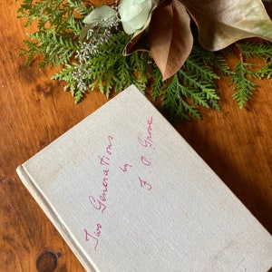 May include: A white book with the title "Two Generations" written in pink ink, followed by "by O. Grove". The book is lying on a wooden surface with green foliage around it.