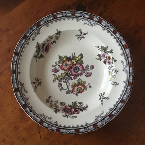 May include: A white ceramic soup bowl with a floral design. The bowl has a blue, black, and red border with a white and gold floral pattern in the centre.