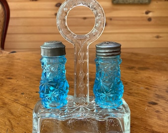 May include: A glass salt and pepper shaker set with a blue glass design. The shakers are on a clear glass stand with a handle.