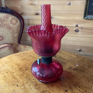 May include: A vintage ruby red glass oil lamp with a round base and a ruffled top. The lamp has a spiral chimney and a metal wick adjuster. The lamp is on a wooden table.
