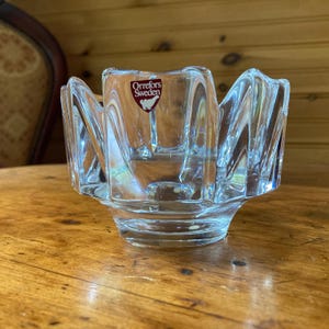 May include: A clear glass candle holder with a distinctive, scalloped design. It features a central cup and a wide, flared base. The words "Orrefors Sweden" are visible. The glass is faceted, creating a play of light and shadow.