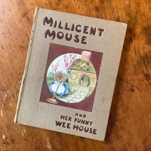 Może przedstawiać: Antyczna książka dla dzieci zatytułowana "Millicent Mouse and Her Funny Wee House". Okładka przedstawia brązową mysz w niebieskiej sukience i białym fartuchu, zamiatającą przed małym, kolorowym domem.