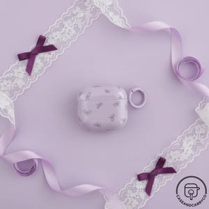 May include: A lilac AirPod case featuring a repeating bow design. The case has a matching lilac ring for attachment. The image is styled with purple ribbon, lace, and bows on a lilac background.