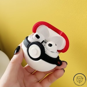 Puede incluir: Funda protectora blanca y roja para auriculares inalámbricos, con diseño de Poké Ball. La funda abierta revela dos auriculares blancos con detalles negros. Fondo amarillo.