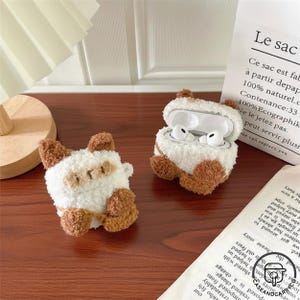 Op de afbeelding: Twee pluche witte en bruine katvormige AirPod-hoesjes. Eén hoesje is gesloten en de andere is open en onthult witte oortelefoons. De hoesjes hebben bruine oren, poten en accenten. De hoesjes staan op een houten oppervlak.