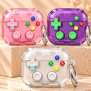 May include: Three protective cases for wireless earbuds, designed to resemble retro game controllers. The cases are transparent in pink, purple, and white, featuring a directional pad, two joysticks, and four action buttons labeled A, B, X, and Y.