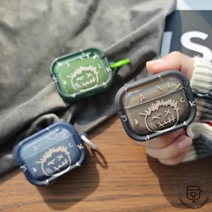 Könnte beinhalten: Drei transparente Schutzhüllen für kabellose Ohrhörer in Grün, Blau und Schwarz. Jede Hülle hat ein Cartoon-Gesicht-Design mit weißen Akzenten und einem silbernen Karabinerhaken. Die Hüllen dienen zum Schutz der Ohrhörer.