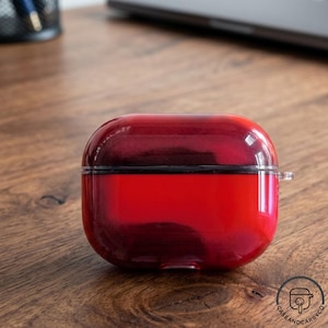 May include: A red and black AirPods Pro case with a glossy finish. The case is rectangular with rounded edges and a black band around the middle. The case is on a wooden surface.