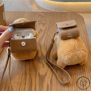 May include: Two tan plush capybara-shaped cases for wireless earbuds. One case is open, revealing white earbuds inside, while the other is closed. Both feature a brown leather-like flap with the text Capybara and a matching wrist strap.