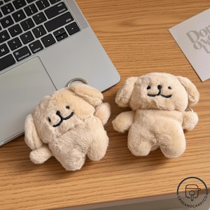 Op de afbeelding: Twee beige sleutelhangers in de vorm van een hond met zwart geborduurde gezichten. Ze liggen op een houten oppervlak naast een laptop en een kleine kaart. De sleutelhangers hebben een zachte, donzige textuur.