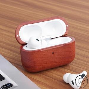 May include: Open wooden case with white earbuds inside and one earbud outside. The case is made of reddish-brown wood with a natural grain. The earbuds are white. The case is on a wooden surface.