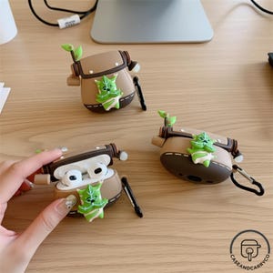 May include: Brown and tan AirPod cases with a green character design. The cases have a small handle and a black carabiner clip. One case is open, revealing white earbuds. The cases are on a wooden surface.