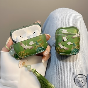 Può includere: Custodia verde per AirPod con design a gru e paesaggio. La custodia presenta un'illustrazione dettagliata di gru, nuvole e montagne in oro e bianco. La custodia è tenuta da una persona che indossa una camicia bianca e jeans blu.