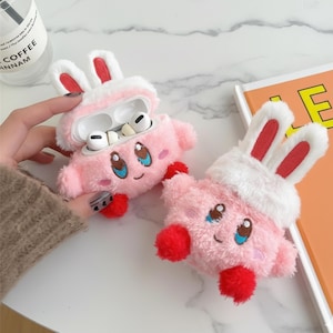 May include: Two pink plush AirPod cases shaped like a cartoon character with bunny ears. The cases have blue eyes, embroidered smiles, and red accents. One case is open, revealing white AirPods.