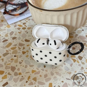 Könnte beinhalten: Weiße kabellose Ohrhörer in einem gepunkteten Etui mit schwarzem Griff. Das Etui ist geöffnet und zeigt die Ohrhörer. Der Hintergrund umfasst eine Kaffeetasse und eine gemusterte Oberfläche.