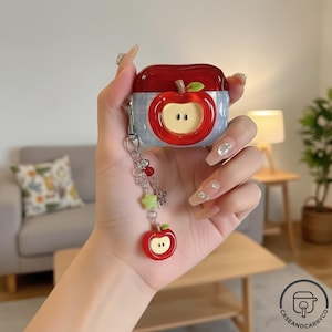 May include: A red and white apple-themed case for wireless earbuds. The case features a red apple design with a green leaf and a silver keychain with charms. The case is held in a hand.
