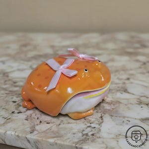 Op de afbeelding: Een keramieken kikkerbeeldje in een levendige oranje kleur met donkerdere oranje stippen, voorzien van een brede mond en een lichtroze satijnen lint gestrikt op de rug. De kikker heeft een witte buik en een gele lijn langs de mond.